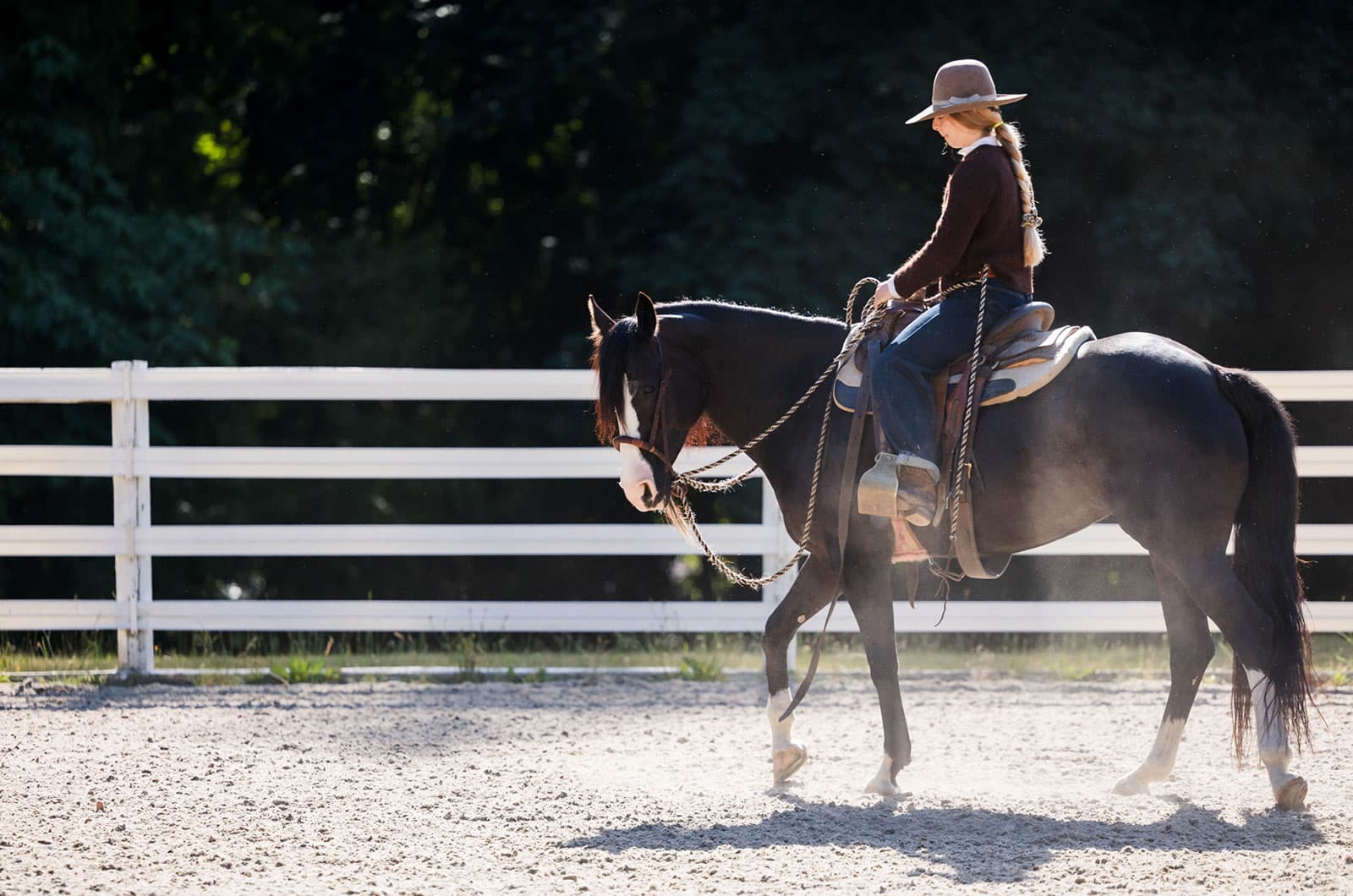 Lili rides along a white rail fence, backlit by the sun